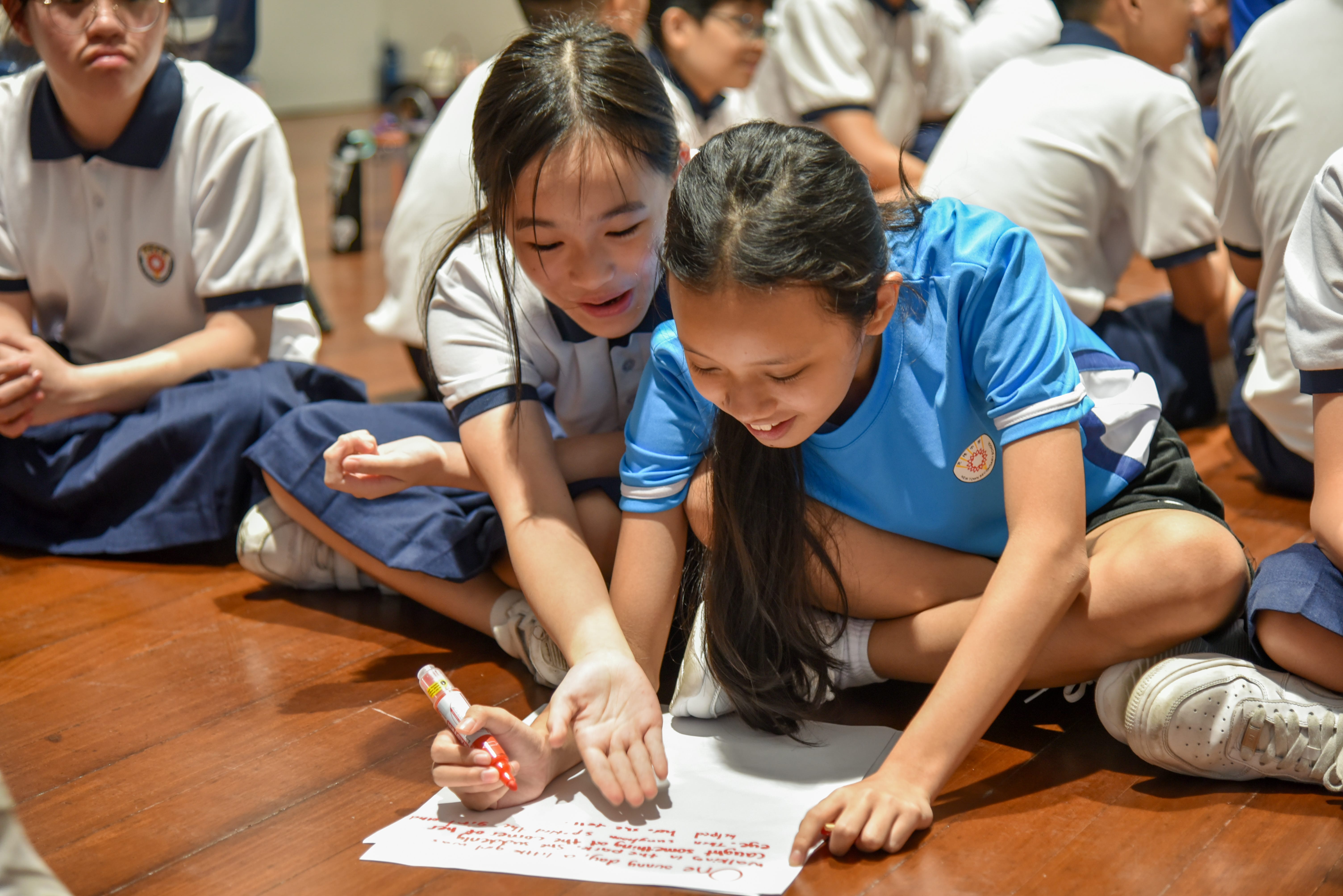 Younger student writing on a piece of paper as another older student instructs her.